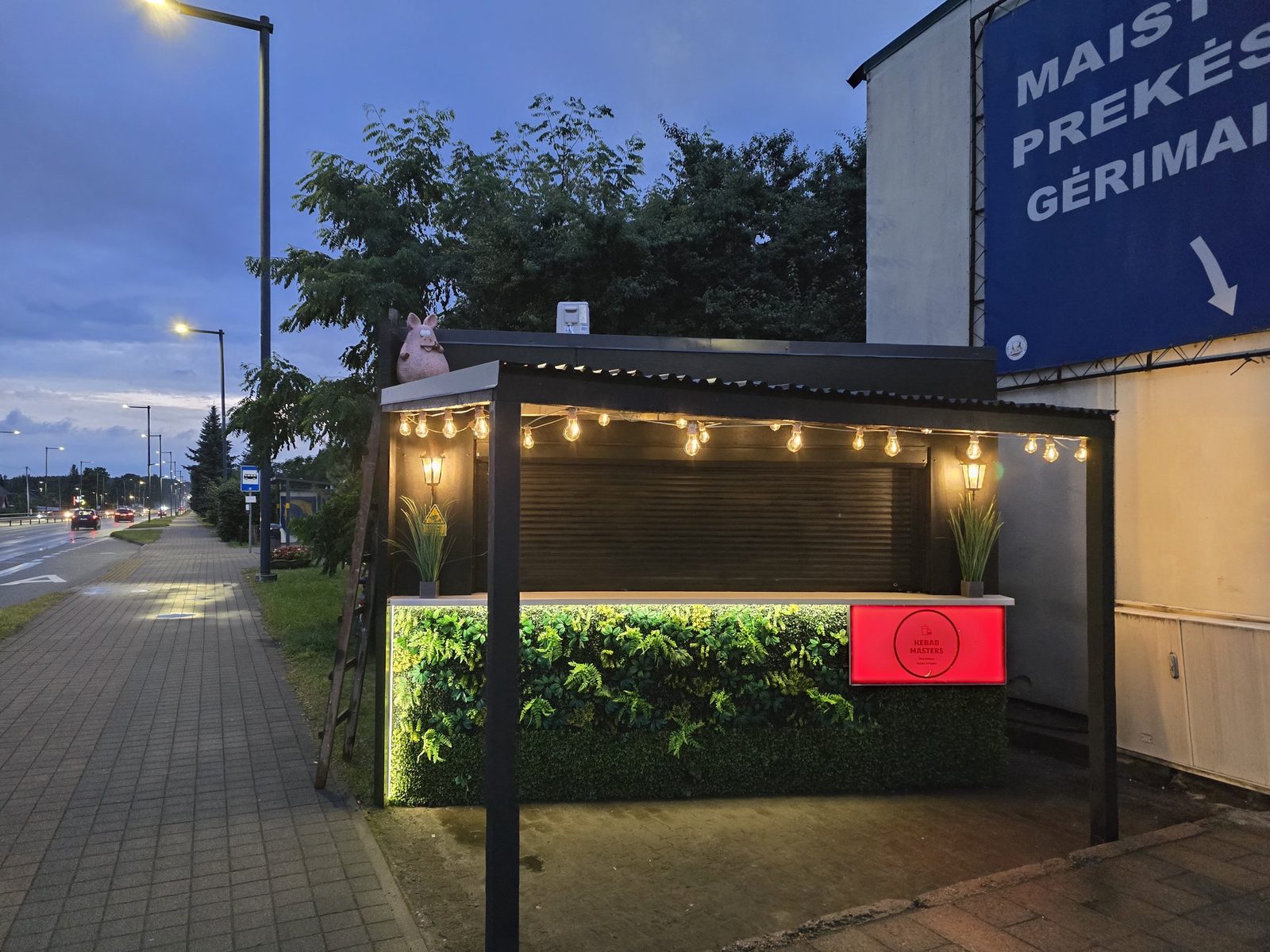 Kebab Masters paviljonas Karmėlavoje vakare: lempučių girlianda, žalia augalų siena ir raudona šviečianti iškaba